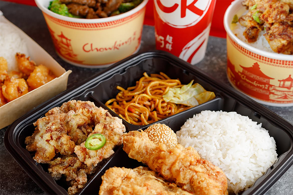 A close-up of a black compartment food tray containing fried chicken, white rice, noodles, and a green jalapeño slice, with Chowking branded takeaway bowls and cups in the background.